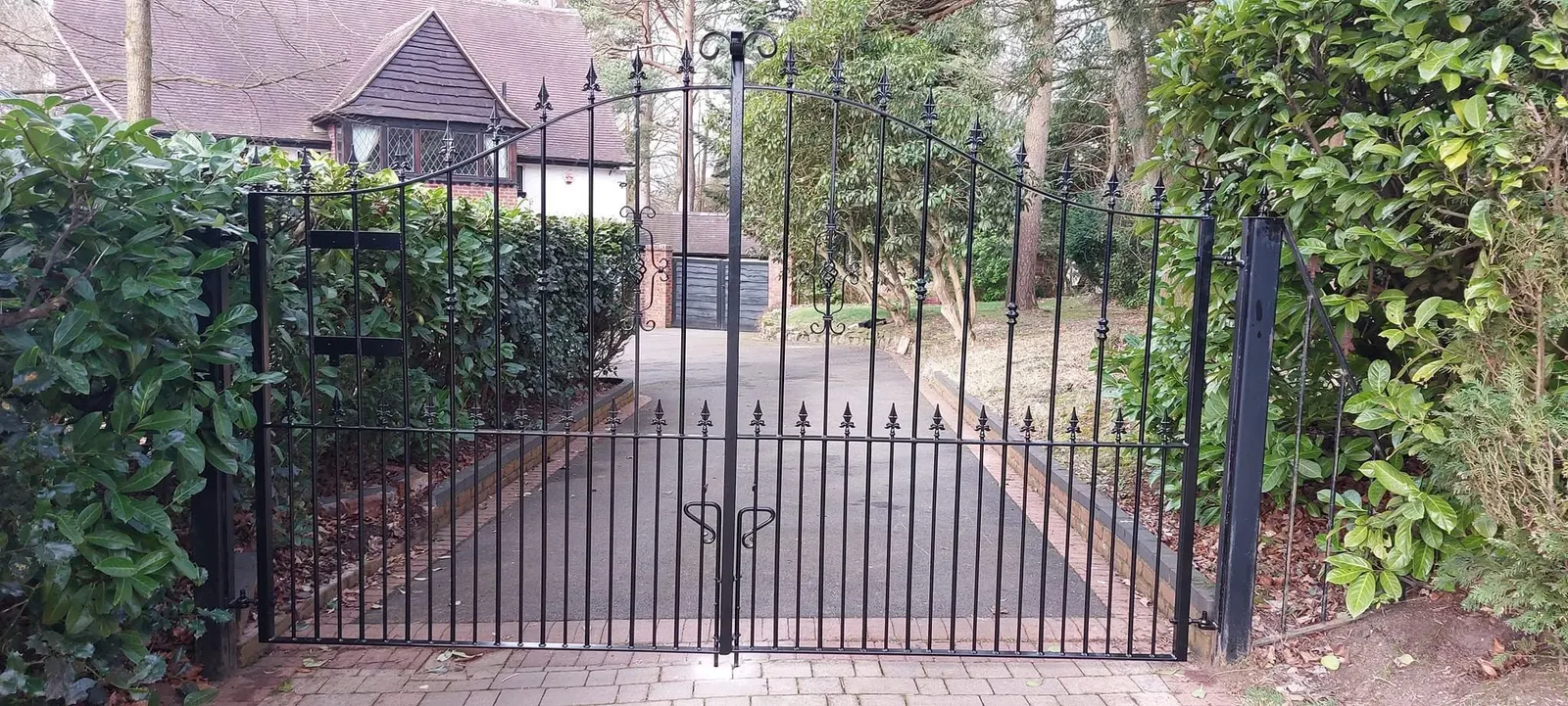 Black arched driveway gates with spear finials in front of a long drive between hedges and trees
