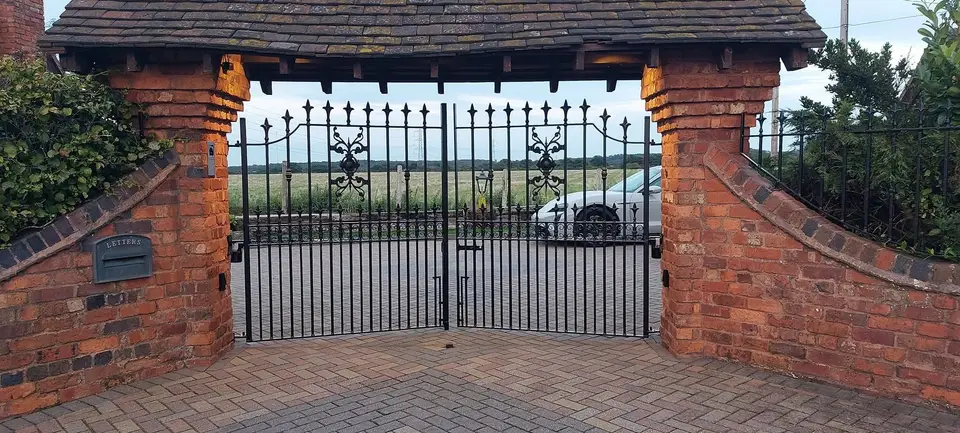 Black arched double gates with scrolled centre panels under a tiled brick archway, open fields beyond