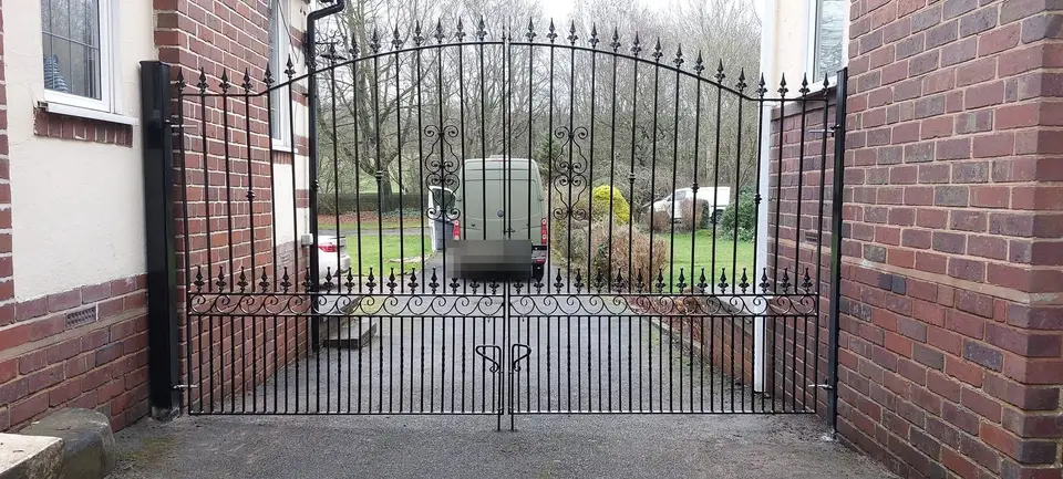 Black arched double gates with spear finials and scrollwork along the bottom rail, fitted between two brick walls