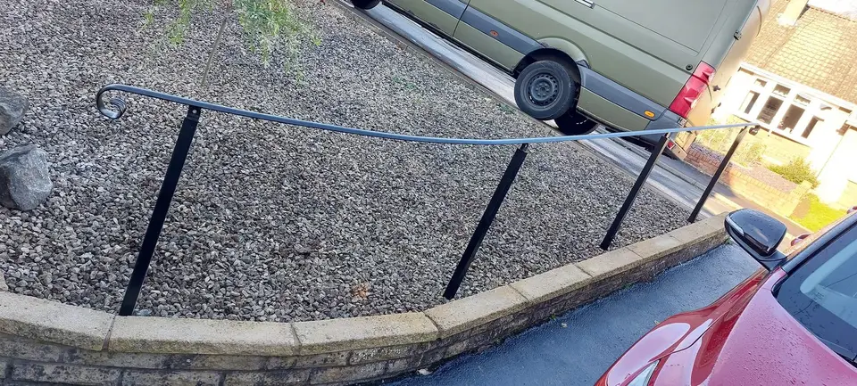 Black curved handrail on raked posts along a low wall at the edge of a gravel driveway