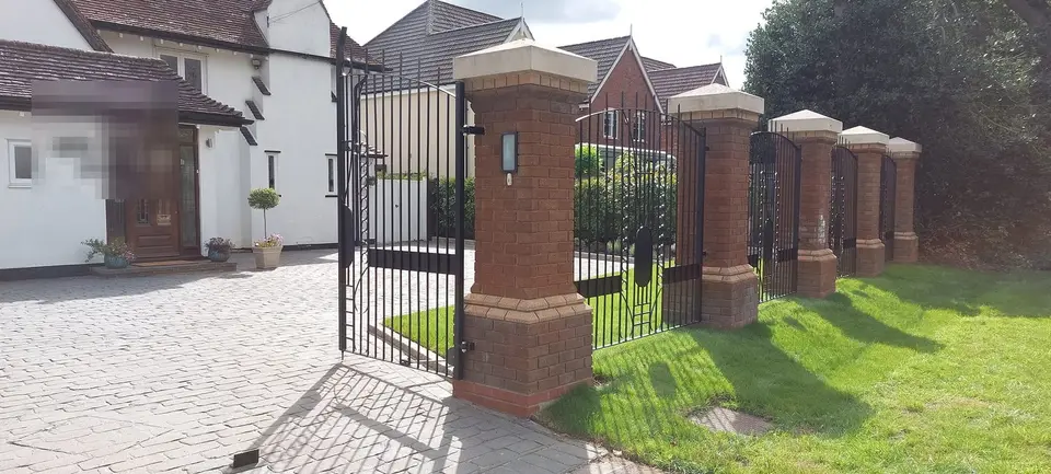 Black driveway gate and railings between tall brick piers with stone caps, on a block-paved drive