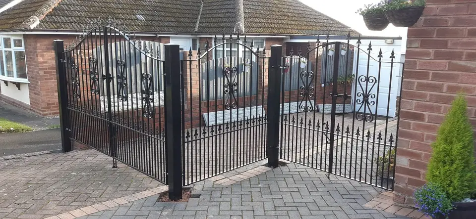Black double driveway gates with spear tops and scroll panels, with tall black posts on a block-paved drive