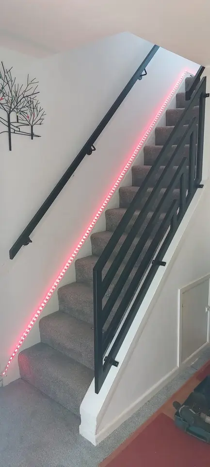 Black wall handrail and bar balustrade beside a carpeted staircase lit by an LED strip