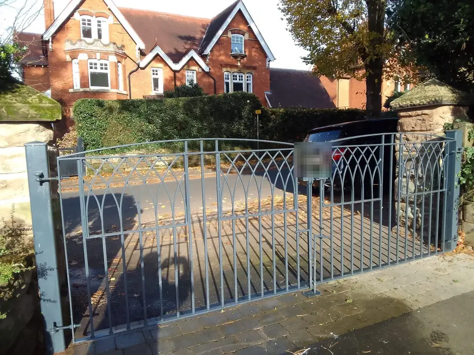 Grey painted steel double driveway gates with a gothic arch pattern, in front of a red brick house with gables