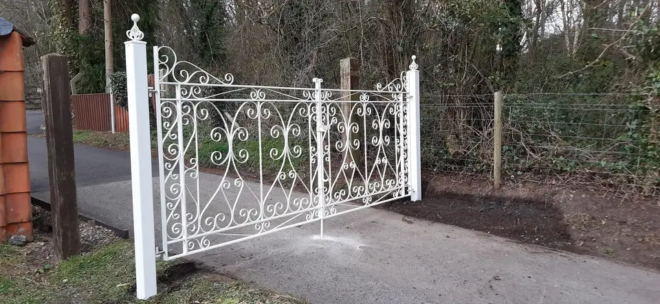 White scrolled wrought iron driveway gate with ball-topped white posts on a tarmac drive in a wooded lane