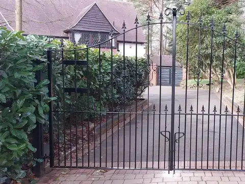 Black arched driveway gates with spear finials in front of a long drive between hedges and trees