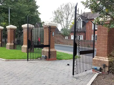 Black driveway gate swung open between brick piers with stone caps, block paving in front
