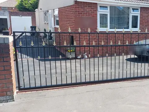 Black bar driveway gates with gold-tipped spear finials across the front of a red brick bungalow