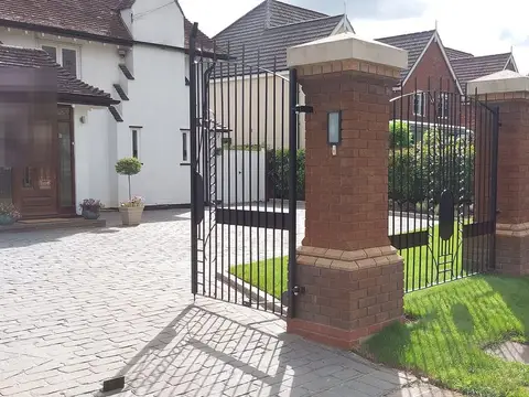 Black driveway gate and railings between tall brick piers with stone caps, on a block-paved drive