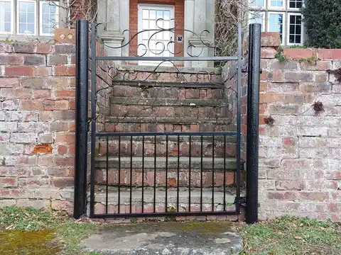 Black pedestrian gate with a scrolled top rail, set into an old brick wall in front of stone steps