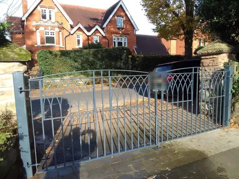 Grey painted steel double driveway gates with a gothic arch pattern, in front of a red brick house with gables